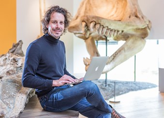 man met laptop in museum met een skelet van een dinosaurus op de achtergrond